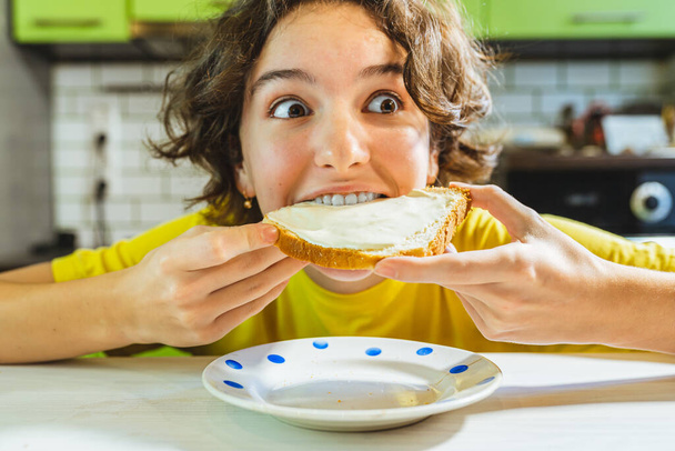 Teenage girl biting cheese sandwich with pleasure, enthusiastic emotion on face. Hungry child eats cheese sandwich with appetite home kitchen, snack lunchtime, junk food, fast food - Photo, Image