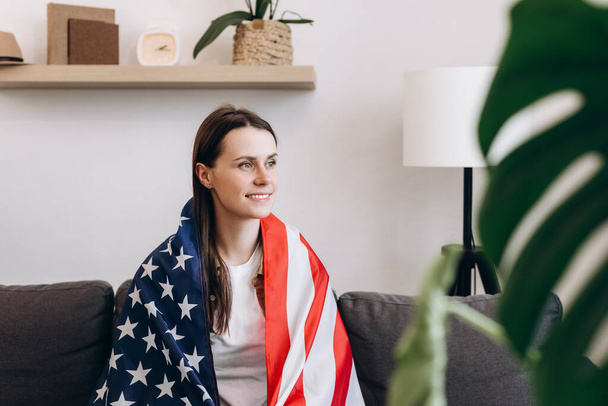 Side view of beautiful pretty lady covered big usa american flag celebrating national traditional holiday, attractive smiling young caucasian woman sitting on cozy sofa at home. Independence concept - Foto, Bild