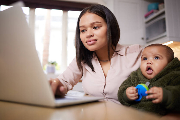 Super Frau macht ihr Ding. Aufnahme einer jungen Frau mit einem Laptop, während sie sich zu Hause um ihr entzückendes Baby kümmert. - Foto, Bild
