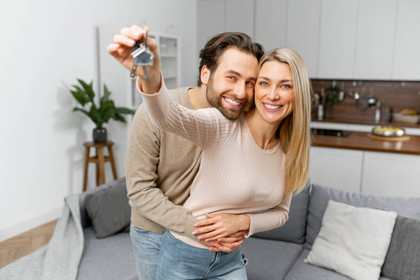 Χαρούμενο καυκάσιο οικογενειακό ζευγάρι που δείχνει τα νέα κλειδιά του σπιτιού της κάμερας ενώ ποζάρει εσωτερικά. Ιδιόκτητη κατοικία, ακίνητη περιουσία και κατοικία - Φωτογραφία, εικόνα