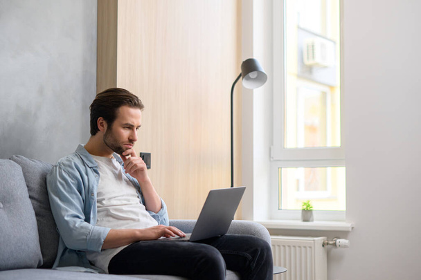 Concentrated caucasian guy looking for issues solving, businessman sitting at the sofa and staring thoughtfully at the laptop screen - Photo, Image