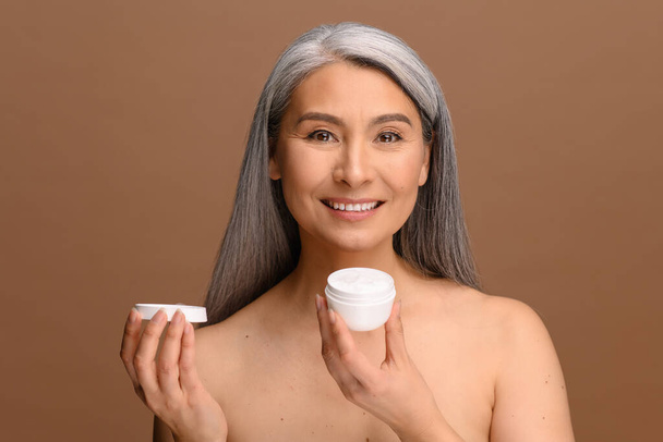 Serene middle-aged Asian woman with fresh clean healthy skin stands with jar of moisturiser anti-aging cream - Φωτογραφία, εικόνα