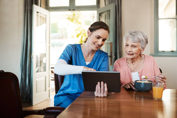 使いやすさをご覧ください。老人ホームでの朝食時にデジタルタブレットを使用している若い看護師と高齢女性のショット. - 写真・画像