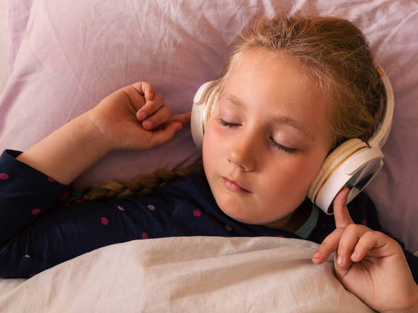 Blondynka dziewczynka w słuchawkach słuchając muzyki dla lepszego snu relaksujący spokojny podcast dla odprężenia ze smartfonem online w łóżku w domu. Książki audio przeciwko bezsenności dla zmęczonych dzieci. - Zdjęcie, obraz