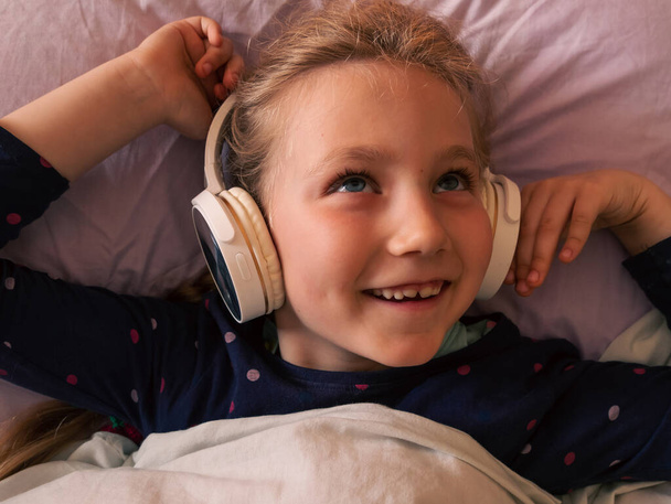 Blonde child girl in headphones learning language listening to music podcast with smartphone online in bed at home. Happy kid listens audio book tales Distance elementary school lesson internet gadget - Photo, image