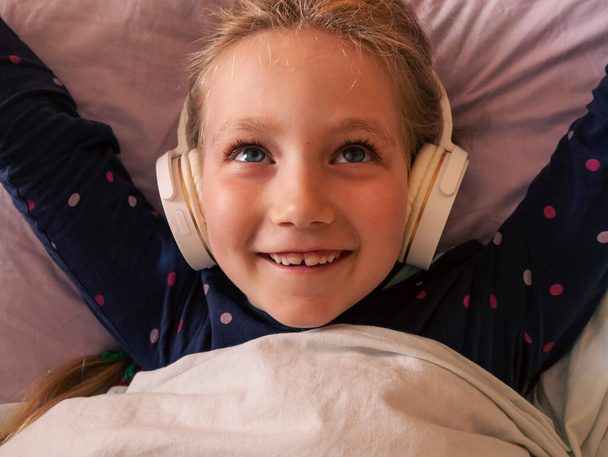 Blond kind meisje in hoofdtelefoon leren taal luisteren naar muziek podcast met smartphone online in bed thuis. Happy kid luistert audio boek verhalen Afstand basisschool les internet gadget - Foto, afbeelding