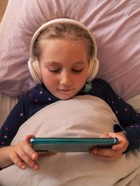 Cute child girl in headphones learning language listening to music audio book podcast smartphone in bed at home. Blogger kid gadget. Technology distance school online lesson chat cell phone conference - Photo, Image