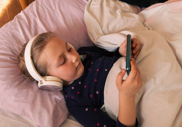 Cute child girl in headphones learning language listening to music audio book podcast smartphone in bed at home. Blogger kid gadget. Technology distance school online lesson chat cell phone conference - Photo, Image