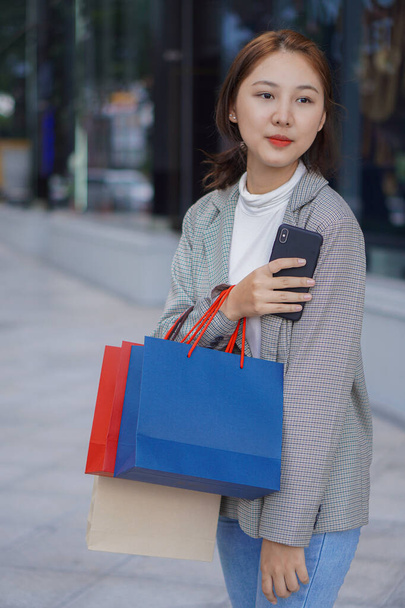 Aziatisch meisje met een tas Gebruik je smartphone tijdens het winkelen in het winkelcentrum. Portret van een mooi meisje dat veel boodschappen doet terwijl ze zich vermaakt na het winkelen op het festival voor het winkelcentrum. - Foto, afbeelding
