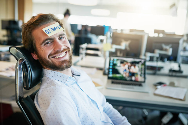 Er ist ein glücklicher Camper. Aufnahme eines fröhlichen jungen Geschäftsmannes mit einem klebrigen Zettel auf der Stirn, der sagt, glücklich zu sein, während er im Büro sitzt. - Foto, Bild