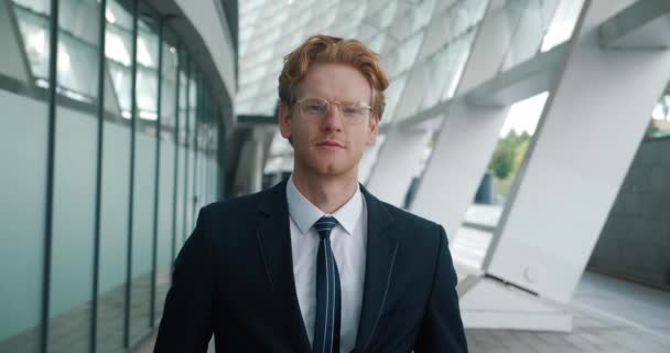 Portrait of ambitious red-haired man top manager looking at the camera and smiling. Young male businessman in formal suitn and eyeglasses standing in the business district near the office building - Filmati, video