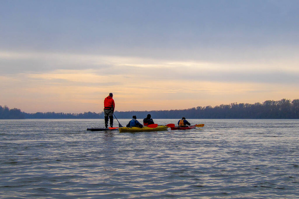 Amigos en kayaks y en stand up paddle board (paddleboard, SUP) remando con una paleta y en el río Danubio invierno en calma fría noche de invierno - Foto, imagen