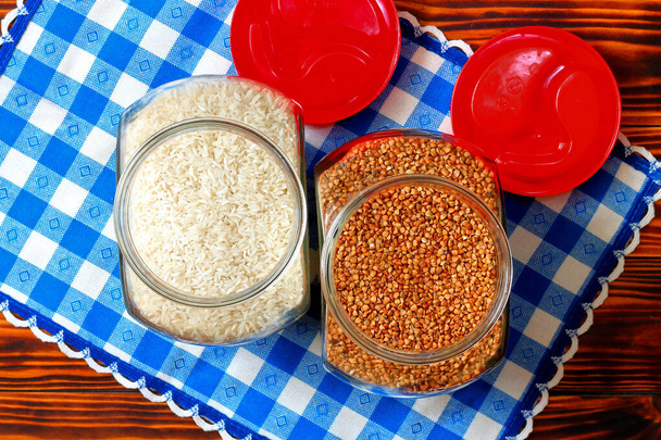 Dwa szklane słoiki z czerwonym wieczkiem z ryżem i gryką na niebiesko-białej serwetce w kratkę. Widok z góry - Zdjęcie, obraz