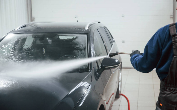 Male worker in uniform washing new modern car indoors. Conception of service. - 写真・画像