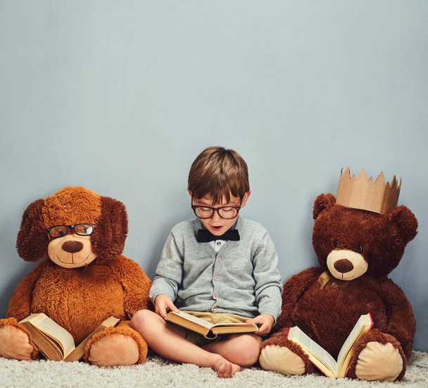 Getting some reading done. Studio shot of a smart little boy reading a book next to his teddy bears against a gray background. - Фото, изображение