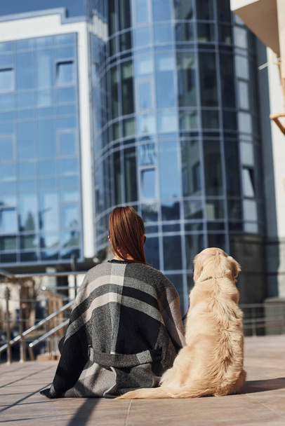 Young positive woman sitting on stairs with her dog when have a walk outdoors near business building. - Photo, image