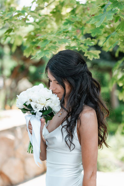 Slanke stijlvolle brunette, gelukkige bruid. Zomer zonnig weer. Botanische tuin met een meer. Witte bloemen - Foto, afbeelding