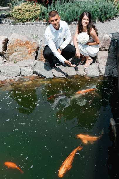 Multicultural happy couple in wedding outfits. The bride has a white bouquet. Botanical garden with a lake. Chinese decorations and fish. Large orange carps. - Foto, Imagen