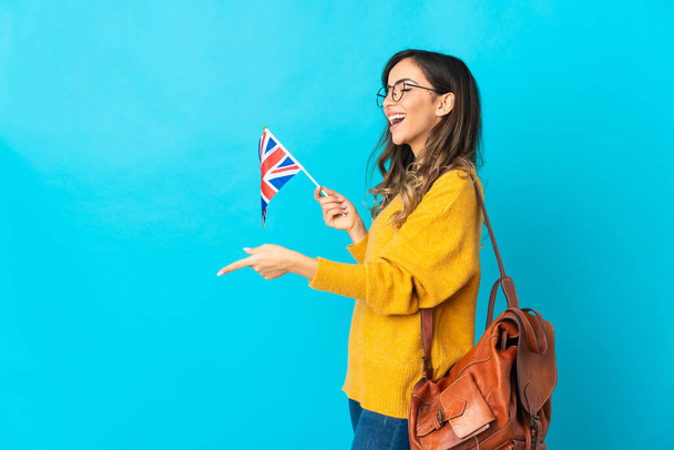 Jeune femme hispanique tenant un drapeau britannique isolé sur fond bleu pointant du doigt sur le côté et présentant un produit - Photo, image