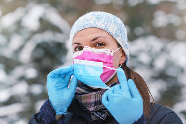 Young woman in warm winter clothing wearing pink disposable virus face mask, putting another one on - some advise that two layers provides better protection again coronavirus covid-19 spread - Photo, Image