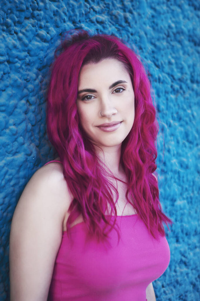 Mladá dívka s růžovými vlasy, opírající se o barevnou stěnu dívající se do kamery. - Fotografie, Obrázek