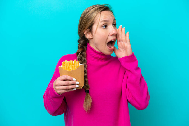 Young caucasian woman holding fried chips isolated on blue background shouting with mouth wide open to the side - Zdjęcie, obraz