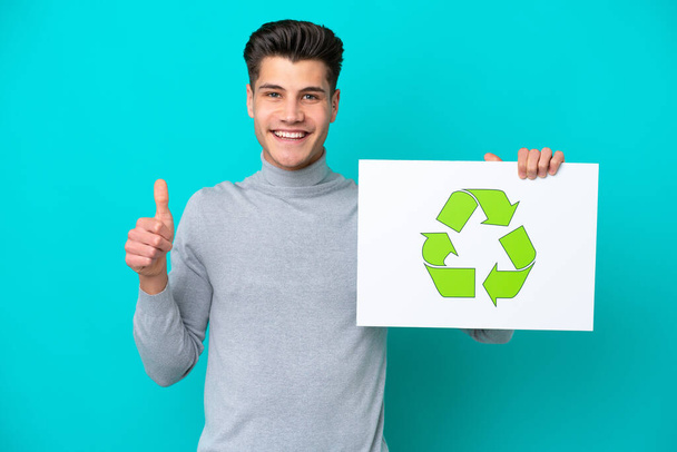 Young handsome caucasian man isolated on blue bakcground holding a placard with recycle icon with thumb up - Foto, immagini