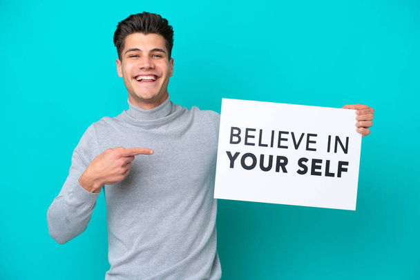 Young handsome caucasian man isolated on blue bakcground holding a placard with text Believe In Your Self and pointing it - Φωτογραφία, εικόνα