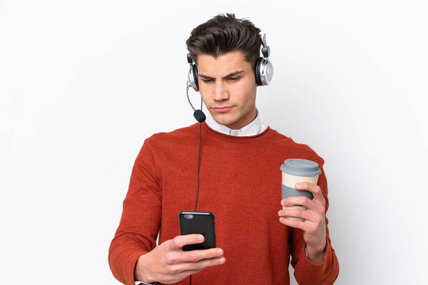 Telemarketer hombre caucásico trabajando con un auricular aislado sobre fondo blanco sosteniendo café para llevar y un móvil - Foto, imagen