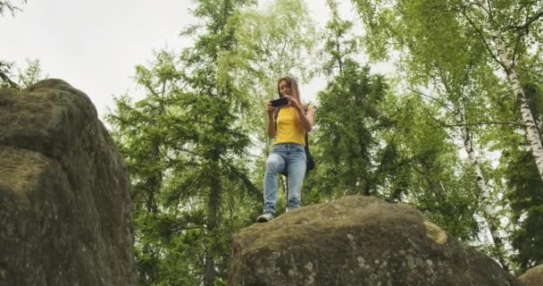 Молодая девушка взобралась на холм и фотографирует в лесу на смартфоне, стоя на камне. Зеленые деревья на заднем плане, вид снизу - Кадры, видео