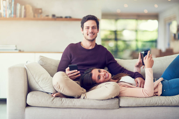 Compartimos nuestras vidas y nuestra radio. Retrato de una joven pareja sonriente usando tabletas digitales mientras se relajan juntos en el sofá en casa. - Foto, imagen