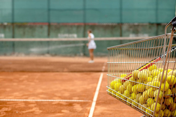 Lots of tennis balls in a metal basket on tennis court on background of tennis player watering the court. - Valokuva, kuva