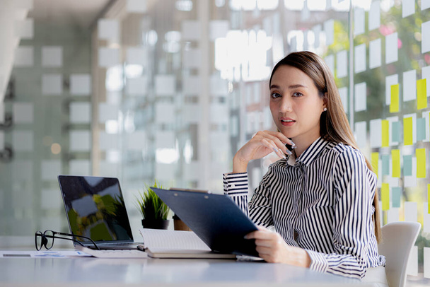 A beautiful Asian businesswoman sitting in her private office, she is checking company financial documents, she is a female executive of a startup company. Concept of financial management. - Photo, Image