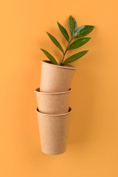 Vertical mockup image of kraft paper coffee cups with green leaves inside - biodegradable, compostable paper utensils for hot drinks. Paper cup on orange background with copy space. Selective focus - Photo, Image
