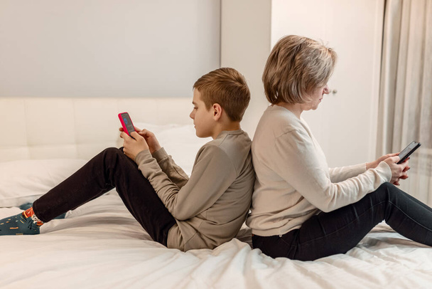 сын и мать-подросток сидят на кровати спина к спине и смотрят на смартфоны, концепцию виртуального общения в социальных сетях, пустую трату времени и оторванность от реального мира - Фото, изображение