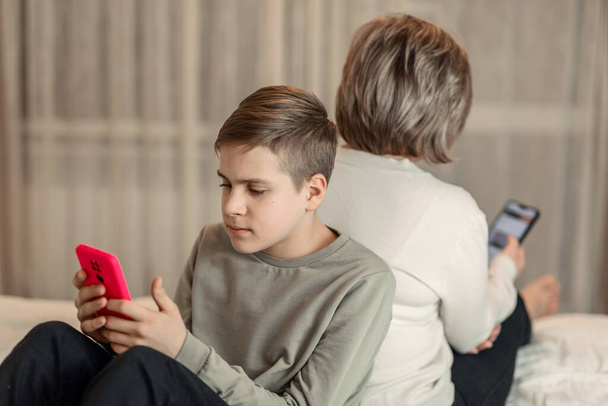 Teenager-Sohn und Mutter, die Rücken an Rücken auf dem Bett sitzen und auf Smartphones schauen, das Konzept der virtuellen Kommunikation in sozialen Netzwerken, Zeitverschwendung und Trennung von der realen Welt - Foto, Bild