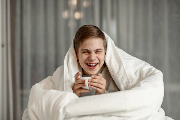 a teenager of European appearance is sitting on the bed wrapped in a blanket with a cup in his hands - Fotó, kép