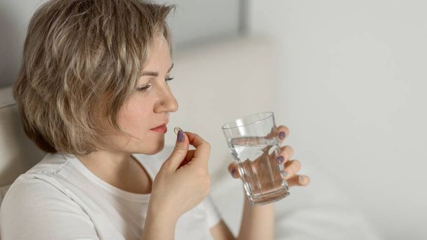 a middle-aged white woman with moisturized beautiful skin holds a glass of clean water and vitamins in her hands while sitting on the bed. the concept of health and beauty care - Photo, image