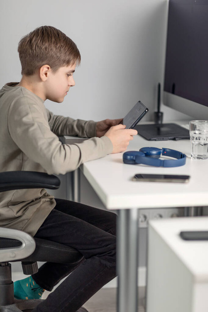 un adolescente biondo si siede a casa a un tavolo davanti a un monitor e partecipa a un incontro online, formazione, giochi per computer - Foto, immagini