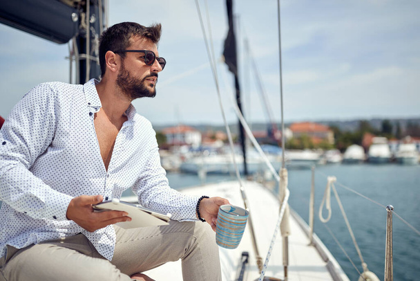 A young man with a tablet is sitting and posing for a photo on the yacht while enjoying a coffee and beautiful scenery of the dock on a sunny day on the seaside. Summer, sea, vacation - Foto, immagini