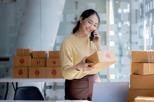 Asian woman is talking on the phone with a customer to confirm an order, she owns an online store, she packs and ships through a private transport company. Online selling and online shopping concepts. - Фото, изображение