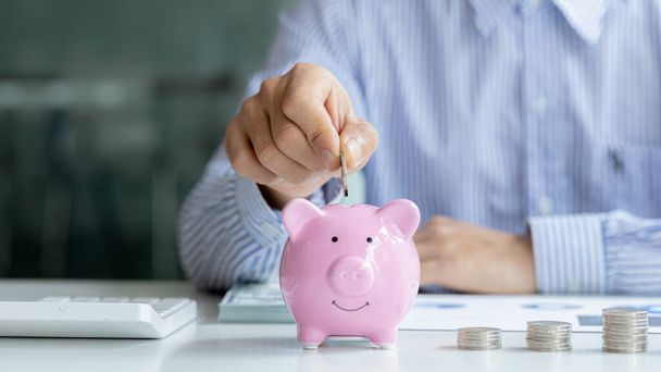 A businessman is putting coins into a pink pig piggy bank, he is saving for investments, money growth, stock investments. The idea of investing money to make it worthwhile to grow and be sustainable. - Photo, Image