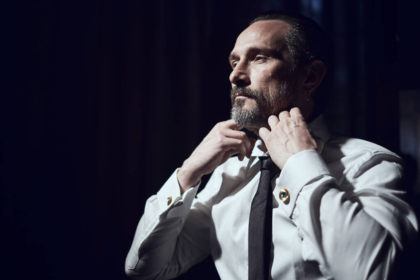 A man puts a black tie on a white shirt in a black studio - Fotoğraf, Görsel