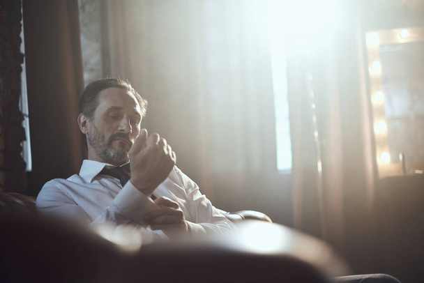A man fastens his sleeves on white shirt sitting on sofa in a smoke room with yellow light - Foto, Imagem
