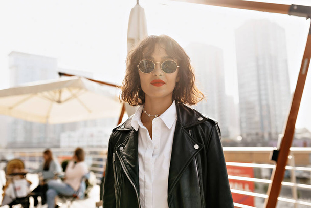 Pleasant young woman in white blouse and black jacket is looking at camera on background of summer cafe. Outdoor photo of debonair girl walking around city in sunglasses. - Foto, Imagen
