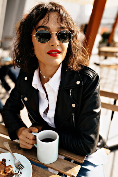 Stylish cute woman with short hairstyle with red lips wearing black jacket and white shirt is drinking coffee outdoor on summer city terrace with happy smile. High quality photo - Фото, изображение