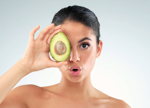 Studioporträt einer schönen jungen Frau, die ihr Auge mit einer Avocado vor grauem Hintergrund bedeckt. - Foto, Bild