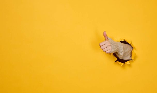 woman's hand sticks out of the torn hole and shows a gesture okay, thumb up. Yellow background - Photo, Image
