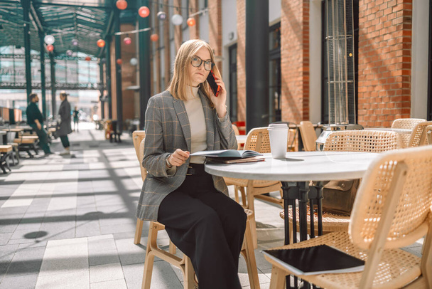 Portrait of young smiling blonde 30s businesswoman calling her best friend, having break, telling something funny, sitting in cafe - Фото, изображение
