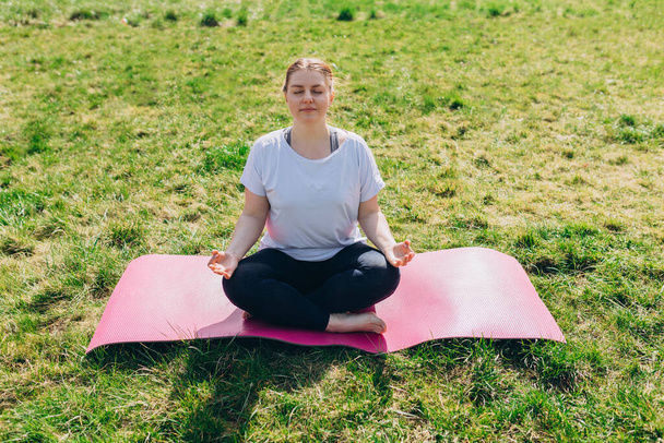 Young smiling attractive redhead woman practicing yoga, girl doing lotus posture. Yoga on grass or sport concept, working out - Photo, Image